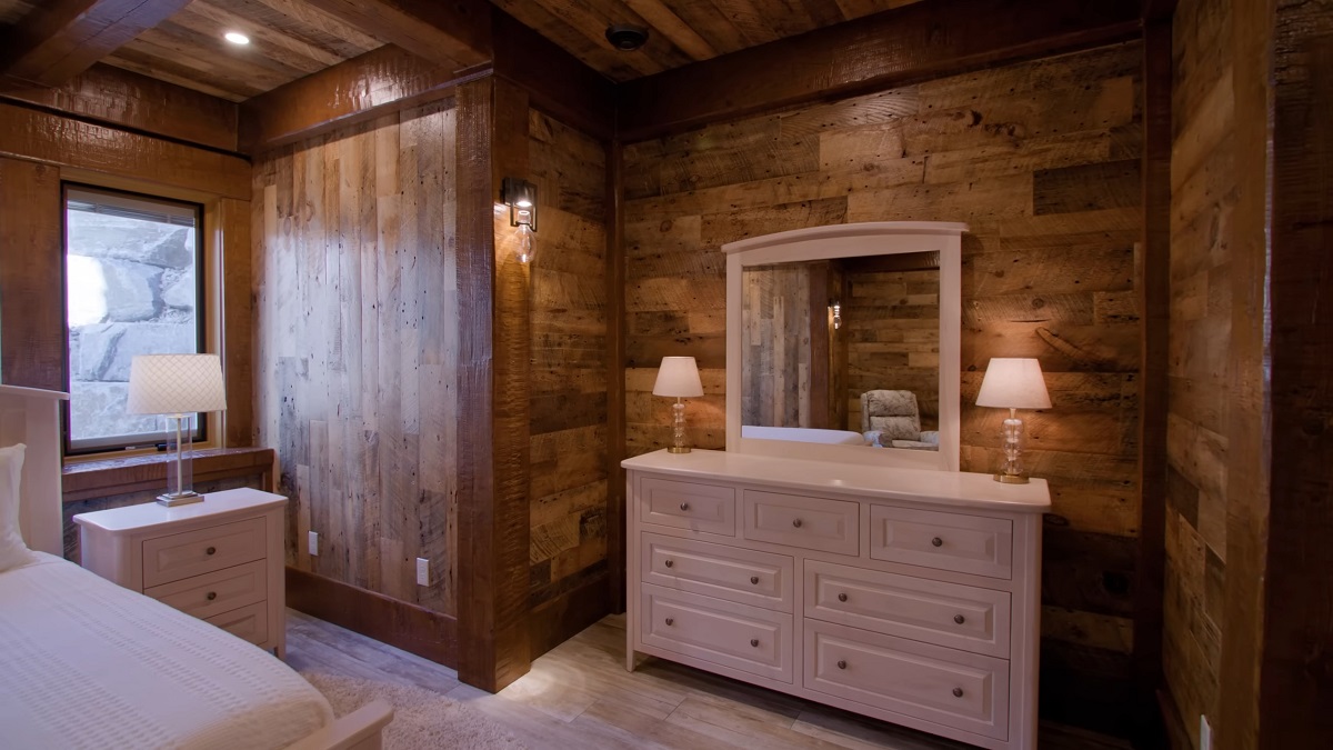 A cozy bedroom with wooden walls, a white dresser, and warm lighting from lamps and wall sconces