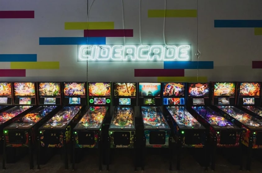 Row of pinball machines under a neon Cidercade sign in a retro arcade venue