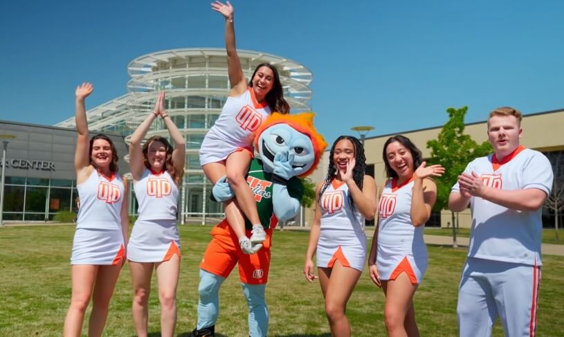 Cheerleaders and mascot pose for a photo in front of a modern building