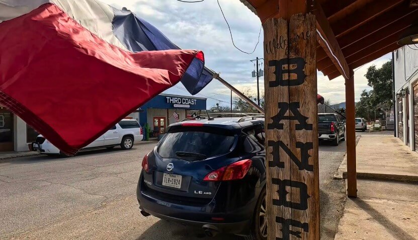 A cloudy day in Texas, with large red, white, and blue flags flapping over a street lined with shops