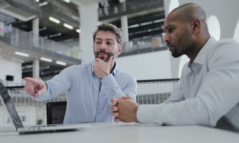 Two men sitting at a table, engaged in discussion while using a laptop
