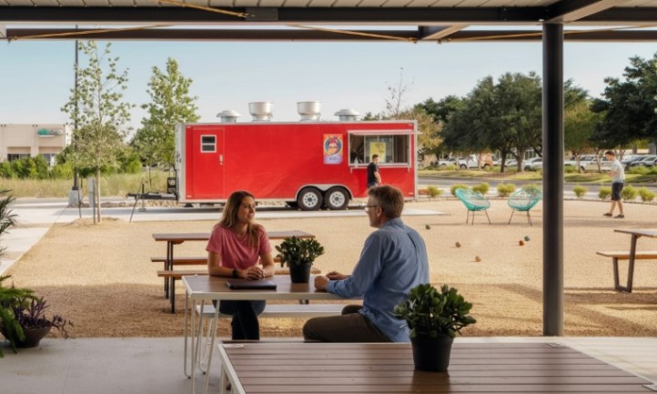 Two people are seated at tables in an outdoor area, enjoying food from a nearby food truck