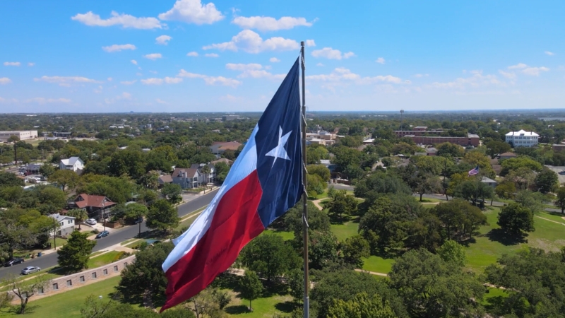 Large Texas flag waves above a city landscape on a sunny day