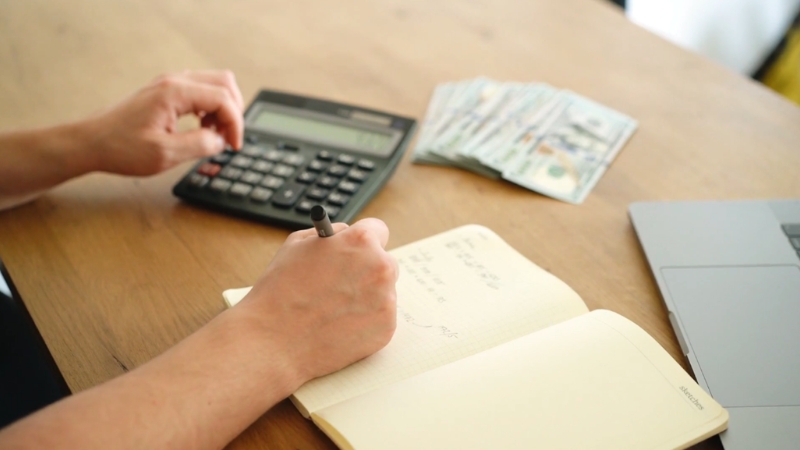 Person calculates expenses with a notebook, calculator, cash and laptop on a desk