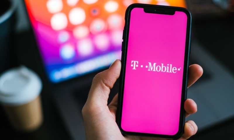 A hand holds a smartphone displaying the T-Mobile logo on a vibrant pink screen