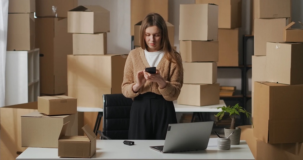 Person managing an online store workspace surrounded by shipping boxes and using a smartphone