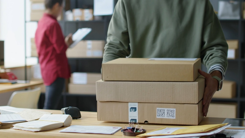 Worker holding packaged orders in a fulfillment space with inventory and paperwork in the background
