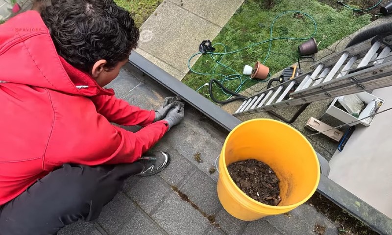 A person in a red jacket, wearing gloves, cleans a rooftop gutter